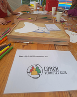 Foto: Zu sehen ist ein Tisch im Innenraum auf dem ein Infoblatt mit der Aufschrift "Herzlich willkommen zu Lorch nernetzt sich" steht, im Hintergrund malen und basteln Kinder.
