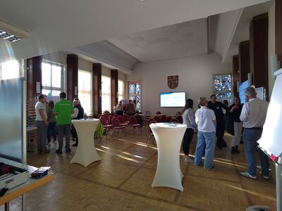 Foto: Zu sehen sind rund 20 Menschen, die im Rathaussaal mit Bestulung zusammen stehen und sich unterhalten.