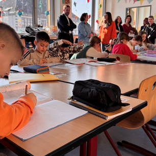 Grundschüler sitzen am Tisch und machen Hausaufgaben, im Hintergrund Pressevertreter und Landrat.