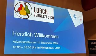 Foto: Zu sehen ist eine Wand, vor ihr ein großer Bildschirm, auf dem eine Powerpointpräsentation abgespielt wird mit dem Titel: "Lorch vernetzt sich - Herzlich willkommen zum Adventstreffen im Dezember 2025 in Lorch".